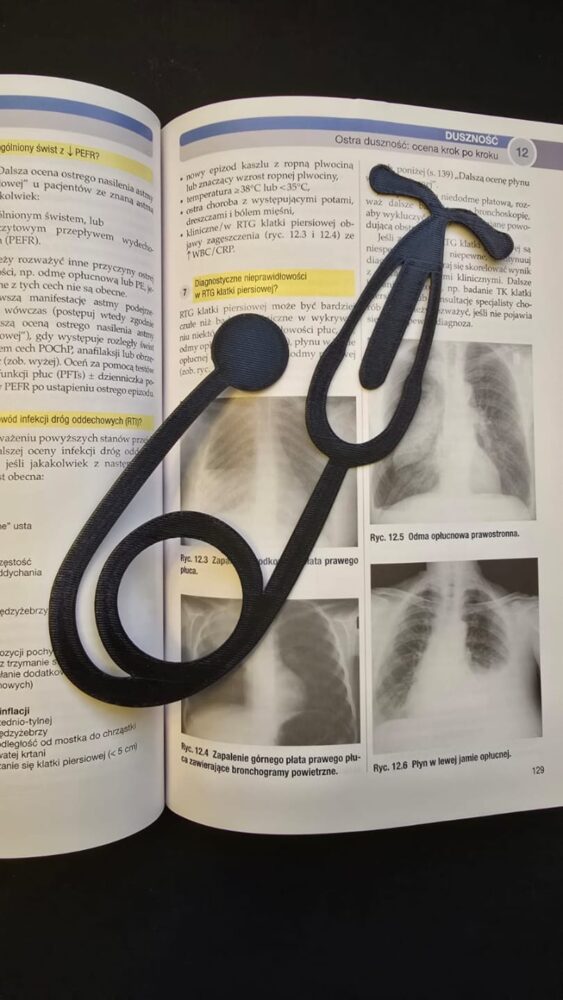 Marcador de página preto em formato de estetoscópio feito em impressão 3D, encaixado sobre a página aberta de um livro de medicina com imagens de raio-X.