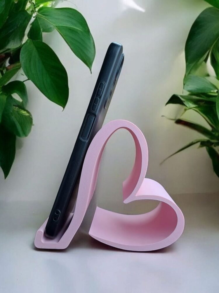 Suporte de mesa para celular na cor rosa claro. O suporte tem um design moderno de fita contínua que forma a silhueta de um coração visto de lado. Um smartphone preto está apoiado nele, com plantas verdes desfocadas ao fundo.