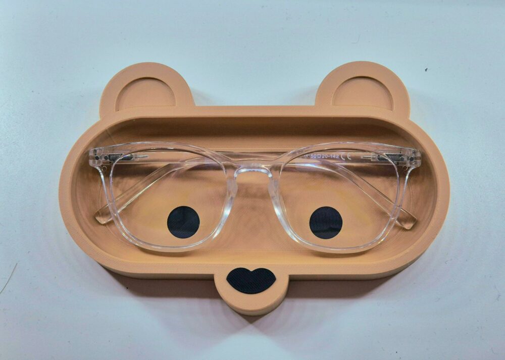 Visão superior de uma bandeja porta-óculos de mesa em formato de rosto de urso na cor bege. O interior possui dois círculos pretos imitando os olhos e um nariz em formato de coração. Um óculos de grau com armação transparente está guardado dentro do recesso do suporte.