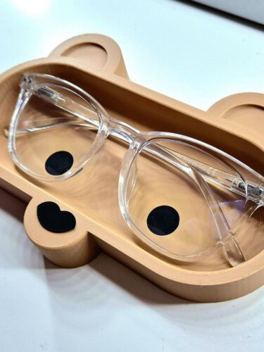 Visão em ângulo de um suporte de mesa bege para óculos com design de ursinho. A textura da peça sugere impressão 3D. Os óculos de armação transparente repousam perfeitamente dentro da cavidade do suporte, protegendo as lentes